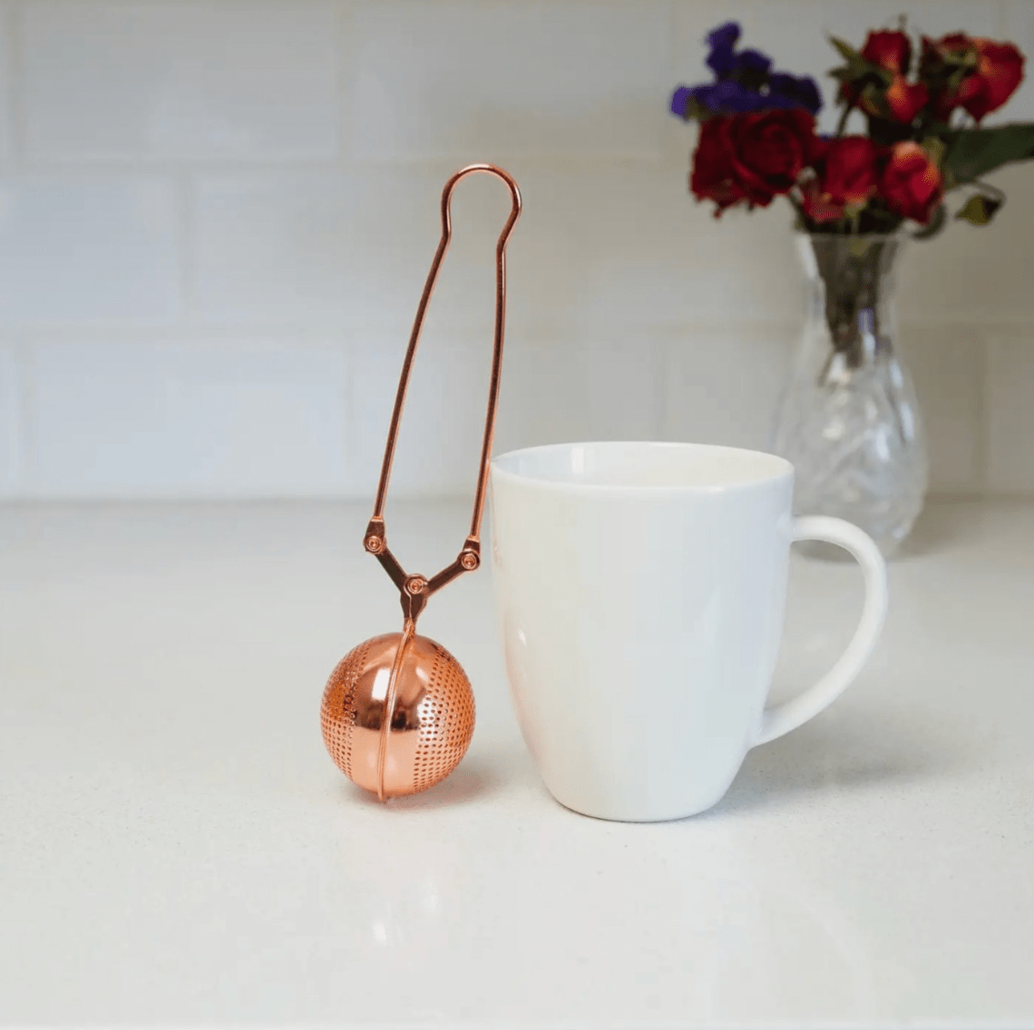 Rose Gold Stainless Steel Tea Strainer | Infuser - marginnotesCopper tea ball infuser next to a white mug on a light surface with flowers in the background.